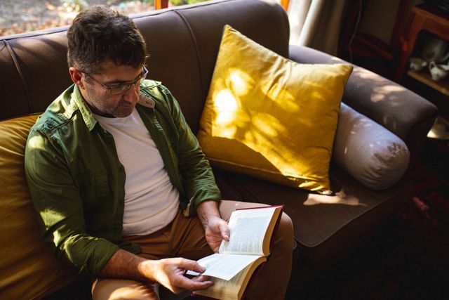 Caucasian man wearing glasses sitting on a sofa and reading a book in a cozy living room. Ideal for use in articles or advertisements related to leisure activities, home lifestyle, relaxation, and personal time. Perfect for illustrating content about reading habits, comfortable home environments, and casual indoor activities.