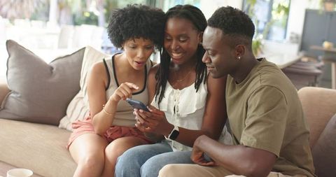 Three friends sharing smartphone on sofa, laughing and smiling in sunlit living room