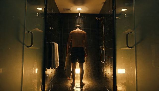 Male showering under ceiling rain shower in moody backlit glass shower stall with towel
