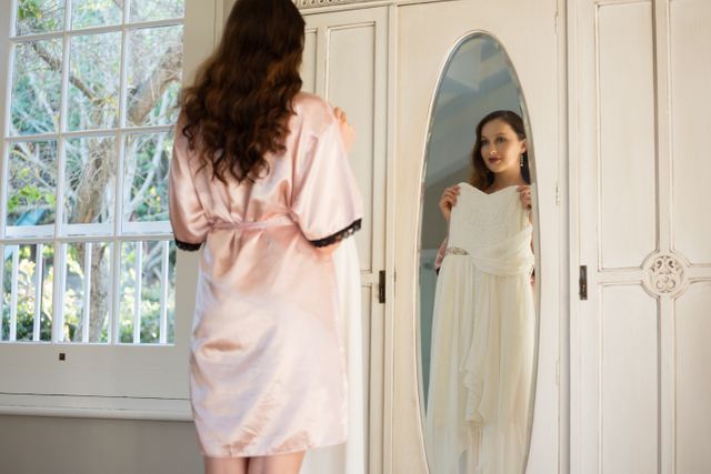 Rear view of bride trying on dress while standing by mirror at home
