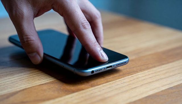 Tapping fingertip on smartphone display on wooden tabletop with metal edge reflection