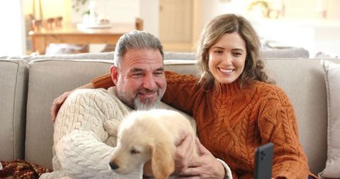 Middle-aged couple cuddling puppy on sofa while video calling on smartphone in cozy home