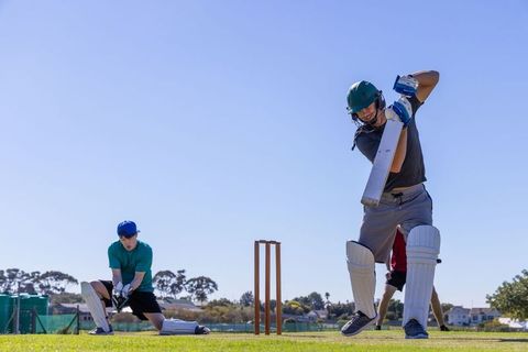 Batsman wearing green helmet and pads swinging cricket bat on sunny suburban pitch