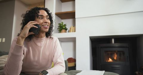 African american nonbinary person talking on phone by fireplace, wearing smartwatch, smiling
