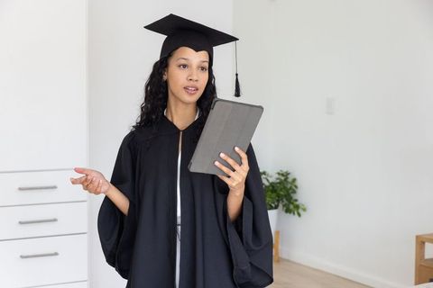 Young graduate in cap and gown holding tablet delivering virtual graduation speech