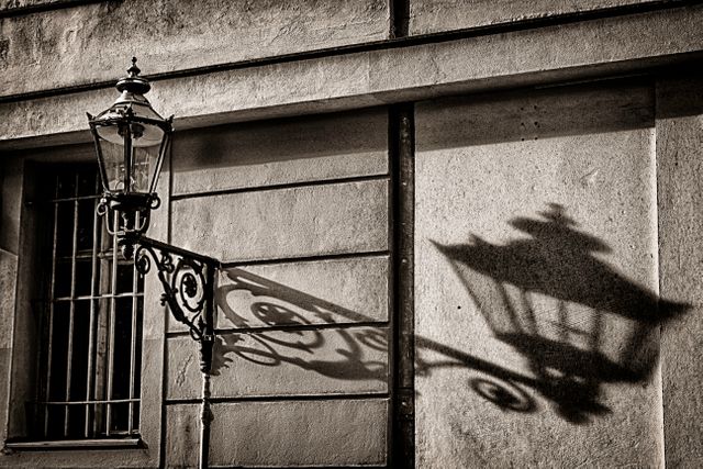 Image shows a vintage wrought iron street lamp casting a detailed shadow on an old building wall in an urban setting. Suitable for use in publications, articles, or blog posts about historical architecture, urban scenery, vintage and antique designs, or city life. Ideal for adding a touch of historical ambiance and architectural detail. Can be used by architectural firms, historians, city planners, or designers.