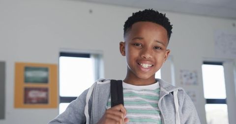Smiling african boy holding backpack strap in bright classroom wearing grey hoodie