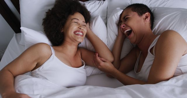 Two women lying in bed under white covers, sharing a moment of laughter. Both wear white sleeveless tops and have their arms intertwined. They look relaxed and happy, enjoying each other's company. This uplifting and intimate scene can be used to depict friendship, bonding, relaxation, love, or morning routines. Ideal for campaigns focusing on happiness, relationships, and self-care.
