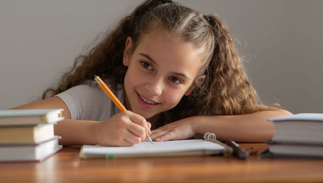 Curly-haired student smiling while writing homework at home desk with textbooks and pencils
