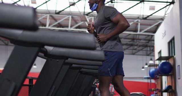 Fit african american man with face mask around his neck running on treadmill in the gym. social distancing quarantine lockdown during coronavirus pandemic