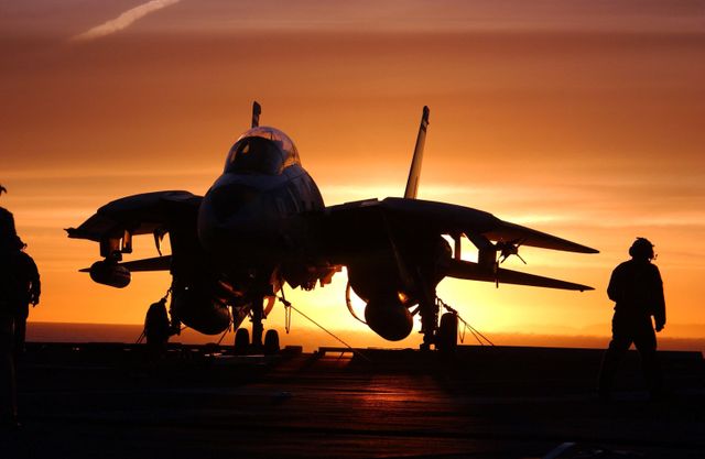 Striking silhouette of a fighter jet on an aircraft carrier deck during sunset. Use this image to highlight military aviation, naval operations, or themes of strength and defense. Also suitable for illustrating articles or promotional materials related to transport, aviation technology, and patriotic themes.