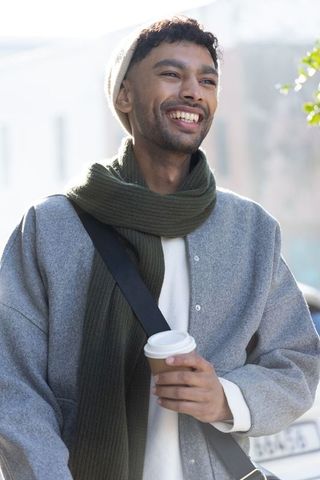 Young asian man smiling with takeaway coffee on sunlit city street wearing scarf and coat