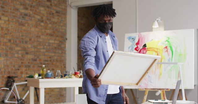 African american male artist wearing face mask using laptop at art studio. art, hobby and creative occupation concept