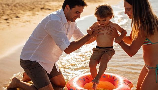 Parents supporting toddler on inflatable donut at sunlit beach during family summer fun