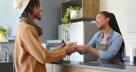 Friendly barista handing coffee to customer at modern rustic cafe with plants and tablet
