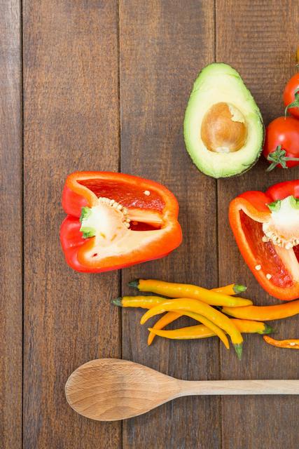 Fresh vegetables including red bell pepper, avocado, yellow chili peppers, and tomatoes arranged on a rustic wooden table with a wooden spoon. Ideal for use in cooking blogs, healthy eating articles, recipe websites, and kitchen decor inspiration.