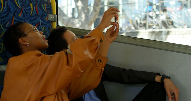 Two friends in casual attire taking a photo together while sitting on a bus during a vacation. The window showcases a glimpse of the city scenery. Ideal for use in travel-related content, tourism promotion, friendship themes, or leisure activities.