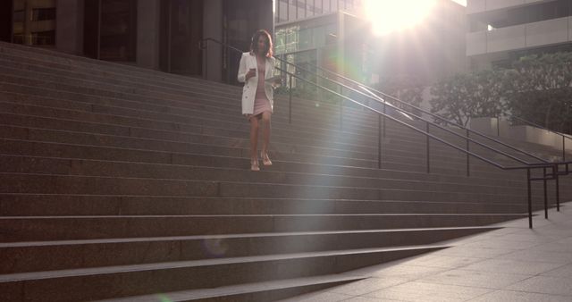 Businesswoman in professional attire walks down stairs in an urban office complex at sunrise with sunlight filtering through. Suitable for use in themes related to business, corporate lifestyle, commuting, urban scenes, and early morning routines.