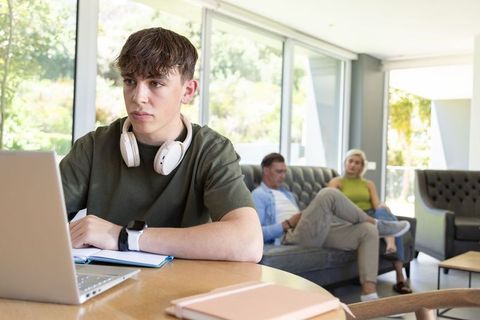 Teenager studying on laptop with headphones in modern living room while parents relax
