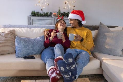 Couple cuddling on sofa wearing santa hat and reindeer antlers, enjoying hot drinks