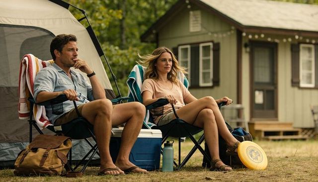 Couple relaxing at campsite near green cabin with cooler, folding chairs and frisbee outdoors