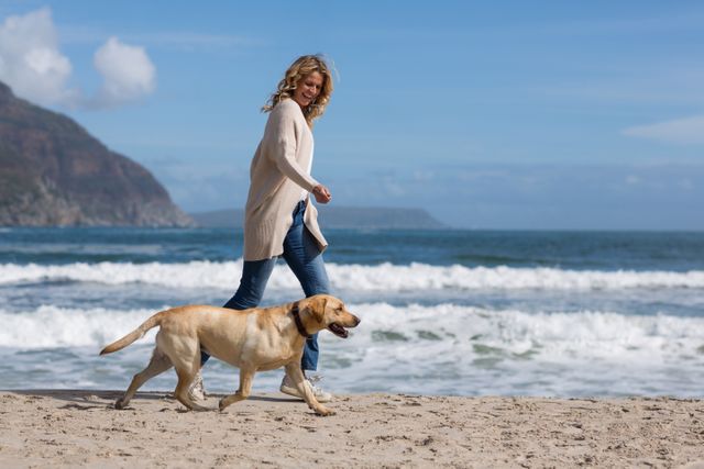Woman walking with her Labrador dog on a sandy beach with ocean waves in the background. Ideal for use in advertisements, travel brochures, pet care promotions, and lifestyle blogs focusing on outdoor activities, relaxation, and pet companionship.