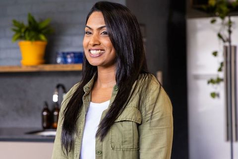 Indian woman smiling in modern kitchen wearing olive green shirt near yellow potted plant