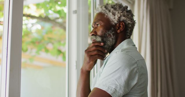 Elderly man with grey hair and beard, dressed casually while looking out a window in deep thought. Captures themes of self-reflection, contemplation, and solitude. Useful for articles or ads focusing on mental health, introspection, retirement, mindfulness, or home settings.