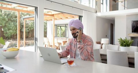 Man in Turban Multitasking with Laptop and Phone in Modern Kitchen