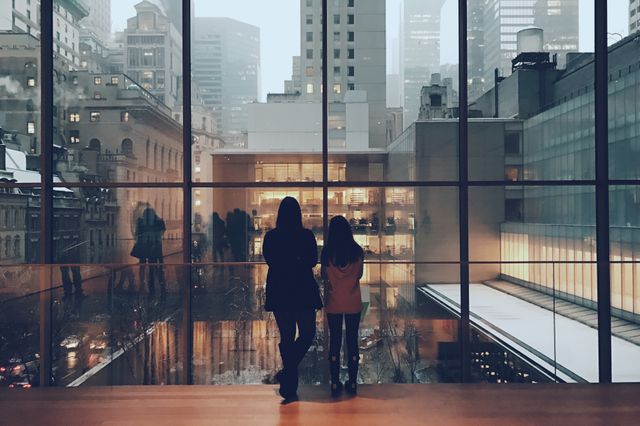 Two individuals stand indoors, looking out through a large glass window at a cityscape featuring tall buildings and skyscrapers. It could be used for themes related to urban lifestyle, modern living, wintertime, architecture, reflection, or observation scenes. The silhouettes against the backdrop of the evening city provide a contemplative mood, suitable for social media posts, promotional materials, or editorial content.