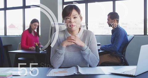 Asian woman clasping hands at desk in modern open office reviewing documents and laptop