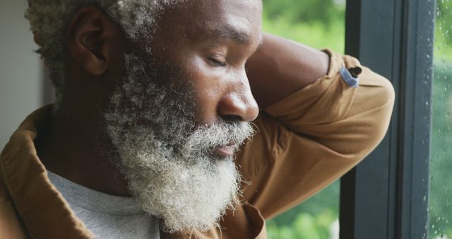 Depicts an elderly man with a beard and silver hair standing by a window, appearing deep in thought. Suitable for use in themes relating to aging, mindfulness, mental health, relaxation, wisdom, and everyday life moments.