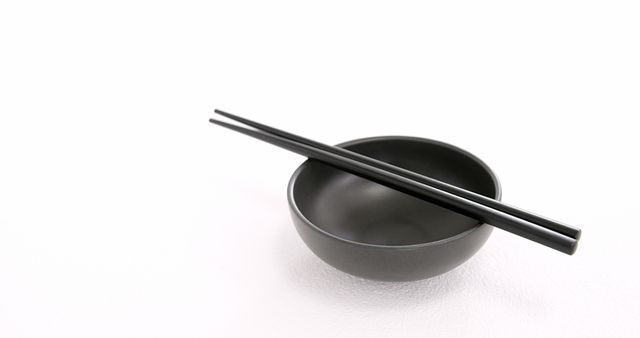 A pair of black chopsticks rests on a matching black bowl, set against a white background, with copy space. This minimalist setup evokes a sense of simplicity and elegance, often associated with Asian dining culture.
