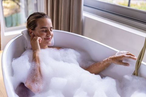 Woman soaking in bubble bath listening to earbuds by window in serene home bathroom
