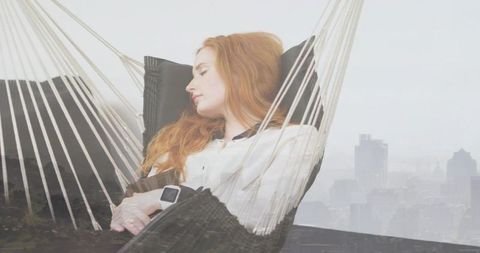Redhead woman relaxing in woven hammock wearing smartwatch with hazy city skyline backdrop