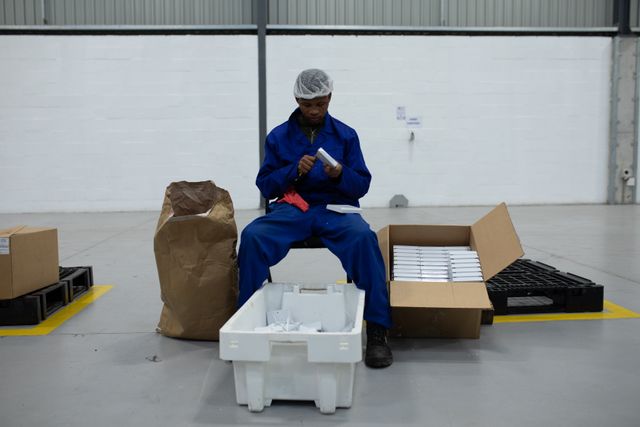 A focused worker is sitting in a factory warehouse, preparing and packing plastic parts into a cardboard box. He is wearing a hair net and overalls, indicating a clean and controlled environment. This image can be used to depict industrial work, manufacturing processes, labor, and production settings.