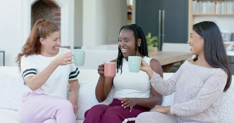 Three diverse women laughing and toasting with mugs on cozy living room sofa