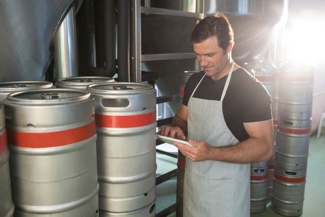 Warehouse worker using digital tablet near metal kegs, ideal for illustrating modern industrial environments, logistics management, inventory control, and technology integration in manufacturing. Useful for business presentations, websites, and articles related to warehousing, brewery operations, and professional work settings.