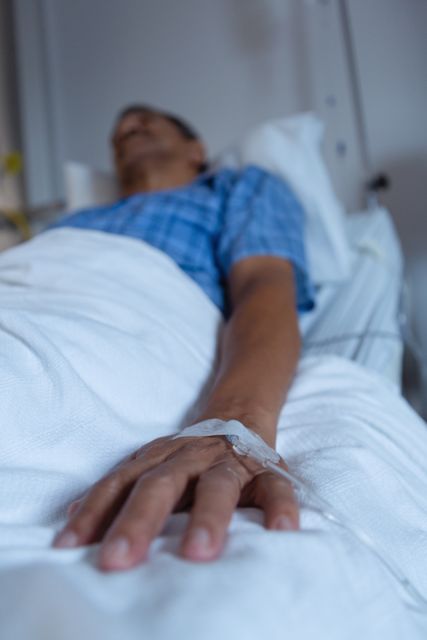 This image shows a patient lying in a hospital bed, receiving treatment through an IV drip. The focus is on the patient's hand and the IV line, indicating medical care and recovery. This image can be used in healthcare-related articles, medical blogs, hospital websites, and educational materials about patient care and medical treatments.