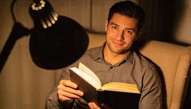Reading man in armchair under warm lamp light holding hardcover book, smiling