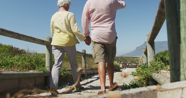 Rear view of senior caucasian couple holding hands and walking on path leading to the beach. travel vacation retirement lifestyle