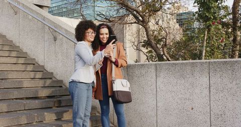 Multicultural women sharing smartphone on urban stairs, smiling friends in casual outfits