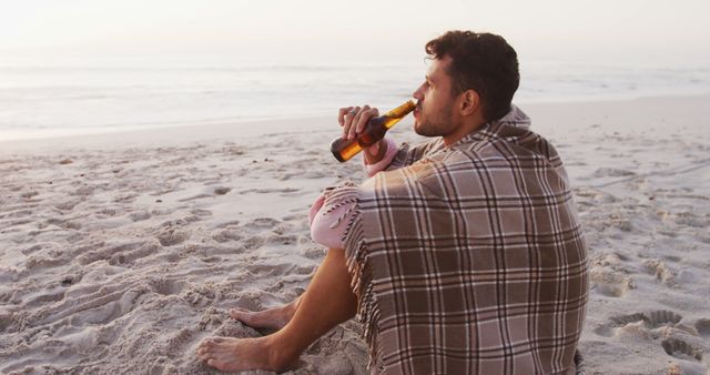Happy biracial man covered with blanket drinking beer on beach. Lifestyle, realxation, nature, free time and vacation.