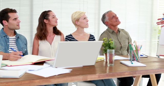 Diverse group of colleagues attentively listening to a presentation at a wooden table with laptop, books, pencils, and notepad. Ideal for illustrating teamwork, corporate discussions, office seminars, and professional training sessions.