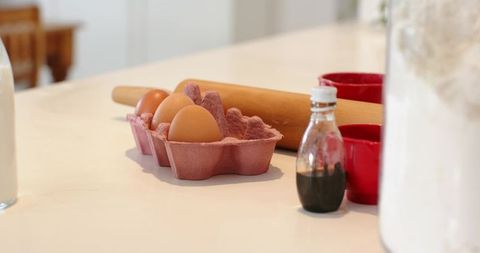 Brown eggs resting beside pink carton, rolling pin, vanilla bottle, flour on white countertop