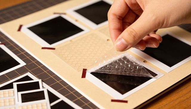 Hand placing protective bubble film over mounted square prints on beige scrapbook page
