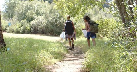 Two women hiking woodland trail carrying backpacks and white bags