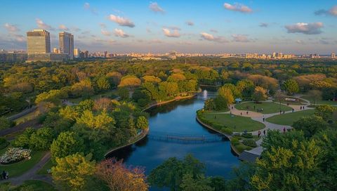 Aerial lush urban park at golden hour showing curving pond, footbridge and distant skyline