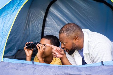 African american father and teen son camping in tent using binoculars bonding outdoors