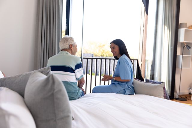 Female doctor in blue scrubs talking with senior man sitting on bed in a home setting. Ideal for use in healthcare, elderly care, home care services, medical consultations, and retirement living promotions.
